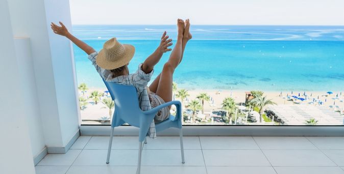 Eine Frau in einem Hemd und einem Strohhut sitzt auf dem Balkon Ihres Hotelzimmers und streckt Arme und Beine in die Luft