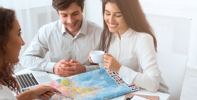 Ein Paar sitzt mit einer Reiseberaterin am Tisch, während die Reiseberaterin ihnen etwas auf einer Karte auf dem Tisch zeigt