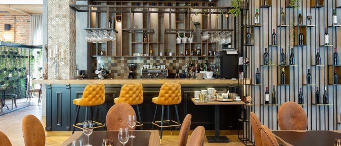 Eine Modern eingerichtete Bar mit gelben Barhockern und einer Wand mit Weinflaschen, im Vordergrund stehen mehrere mit Gläsern gedeckte Tische