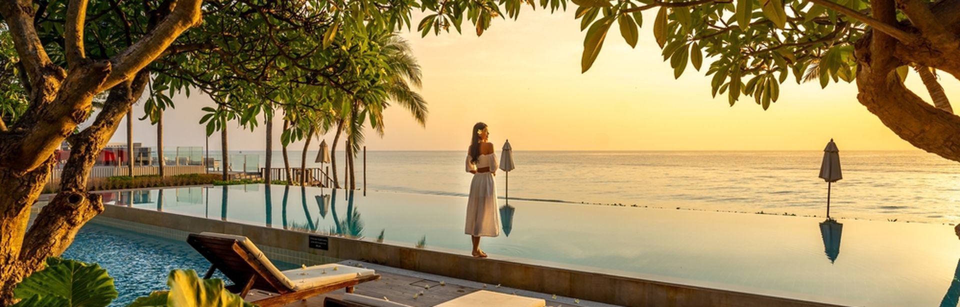 Frau steht bei Sonnenuntergang vor einem Pool der direkt vor einem Meer liegt