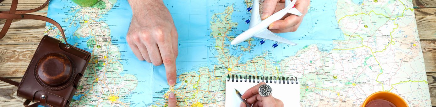 Weltkarte auf Holztisch ausgebreitet, Zwei Personen zeigen mit Fingern auf verschiedene Orte auf der Karte, Eine Person hält ein Modellflugzeug, Notizblock mit Stift, Zwei Tassen stehen auf dem Tisch, Alte Kamera mit Ledertasche