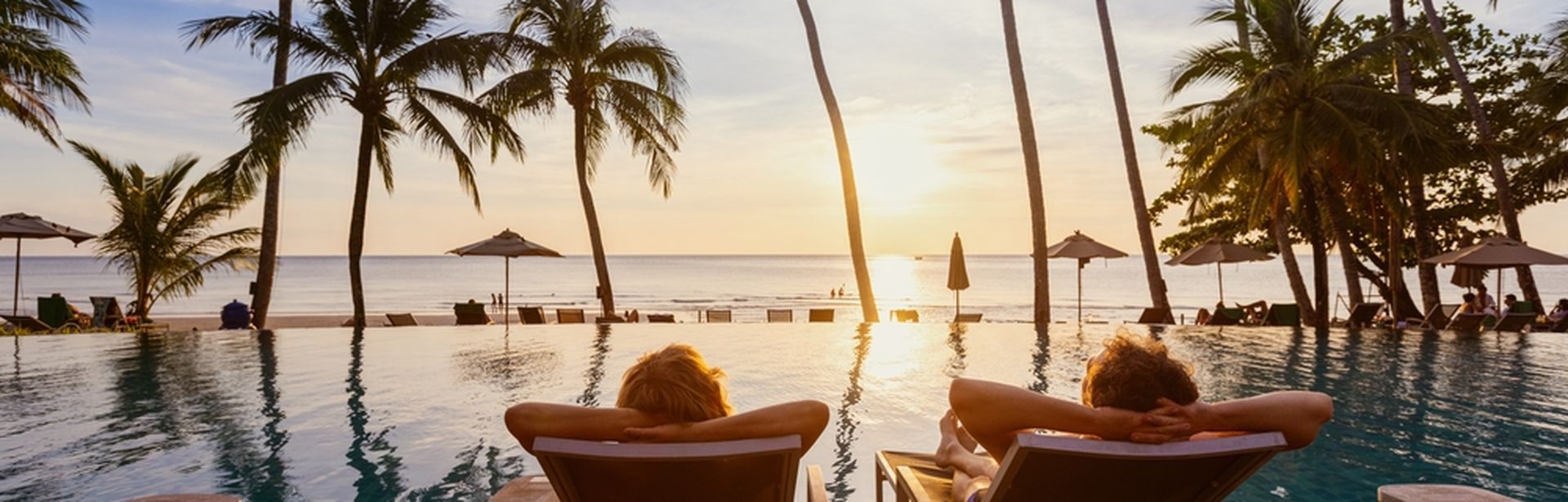 Zwei Personen von hinten fotografiert; Sie liegen auf zwei Liegen auf einem Steg im Pool und schauen auf den Sonnenuntergang über dem Meer