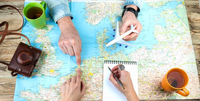 Weltkarte auf Holztisch ausgebreitet, Zwei Personen zeigen mit Fingern auf verschiedene Orte auf der Karte, Eine Person hält ein Modellflugzeug, Notizblock mit Stift, Zwei Tassen stehen auf dem Tisch, Alte Kamera mit Ledertasche