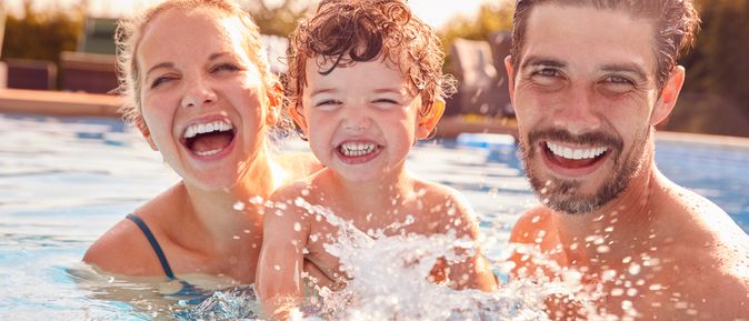 Eltern mit einem Kind spielen im Pool und lachen in die Kamera