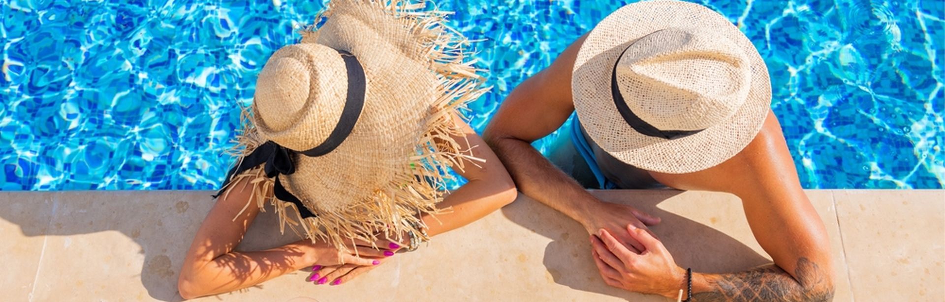 Zwei Personen von oben fotografiert, Sie tragen Strohhüte und man sieht Ihre Gesichter nicht. Sie sind in einem Pool und lehnen am Rand, vor Ihnen stehen Getränke auf dem Boden