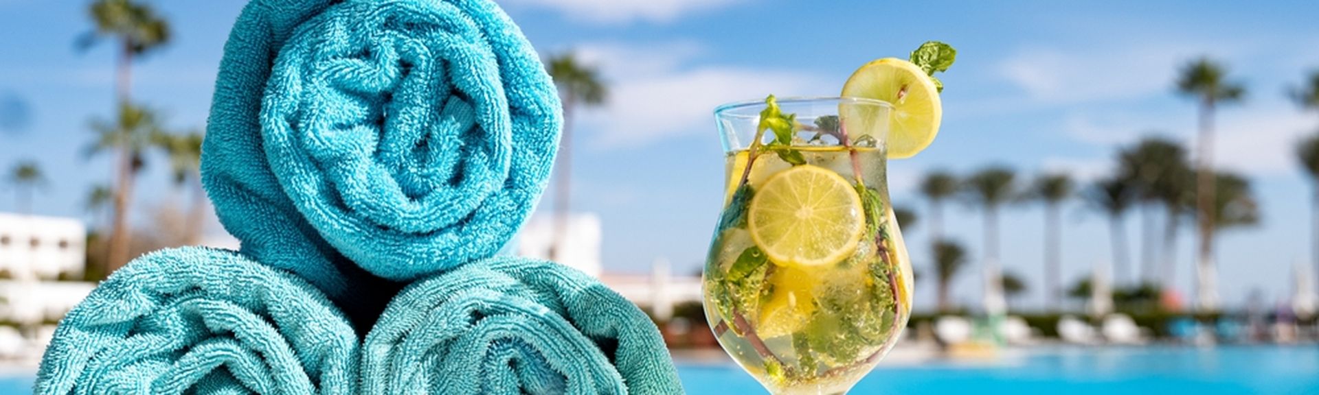 Drei blaue aufgerollte Handtücher liegen am Poolrand, daneben ein gelbes Getränk mit Zitrone