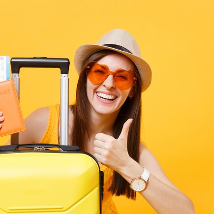 Frau vor gelbem Hintergrund, die hinter einen Koffer hervor schaut und einen Reisepass mit Flugtickets in einer Hand hält. Die Frau zeigt einen Daumen nach oben mit der anderen Hand