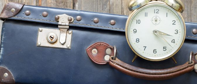 Auf einer Holzoberfläche liegt ein alt aussehender blauer Koffer und ein großer Wecker