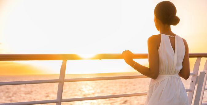 Eine Frau steht an der Reling eines Kreuzfahrtschiffes und schaut in den Sonnenuntergang