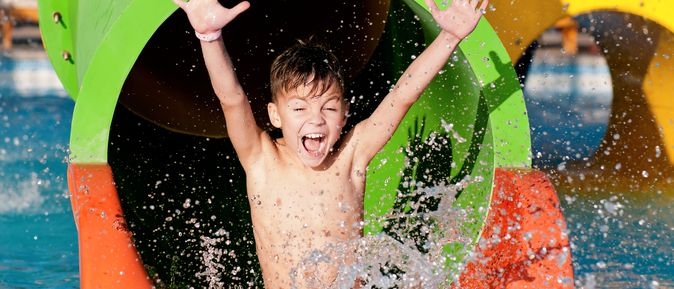 Ein Junge rutscht aus einer Wasserrutsche in einen Pool und reißt die Arme hoch