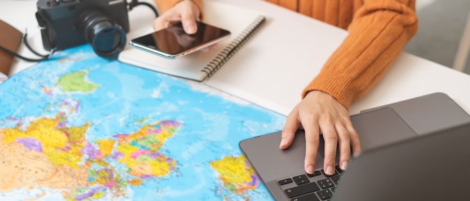 Ein Schreibtisch mit einer Weltkarte als Unterlage, einem Sonnenhut und einer Kamera, eine Person bedient mit der einen Hand ein Handy und mit der anderen einen Laptop