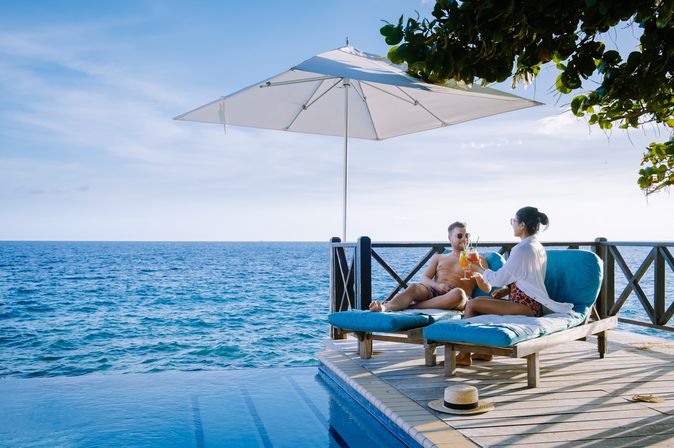 Eine Frau und ein Mann sitzen unter einem Sonnenschirm auf Liegen auf einem Steg und stoßen mit Cocktails an