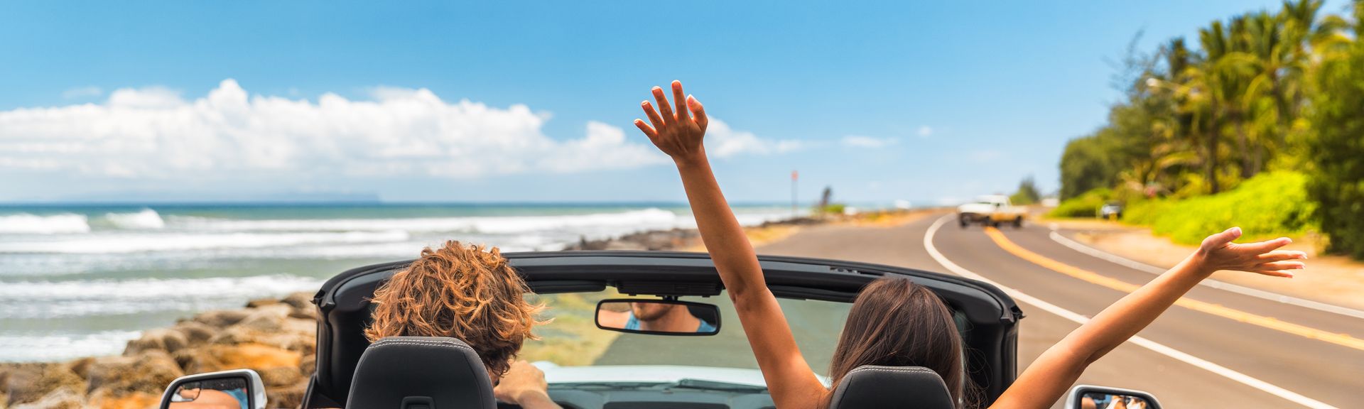 Zwei Personen im Auto mit offenem Verdeck, im Hintergrund kann man das Meer sehen