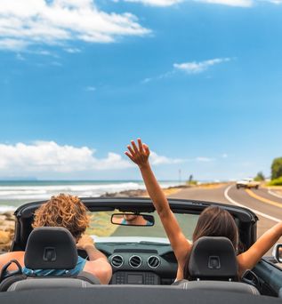 Zwei Personen fahren in einem Auto eine Straße am Meer entlang, eine von ihnen reißt die Arme nach oben