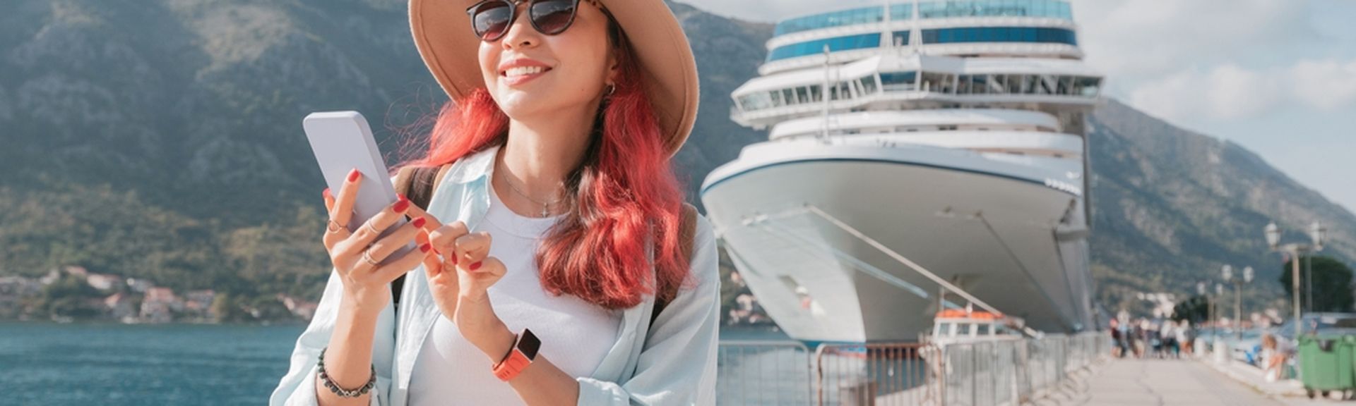 Eine Frau mit roten Haaren steht an einem Hafen, hält ihr Smartphone fest und lächelt