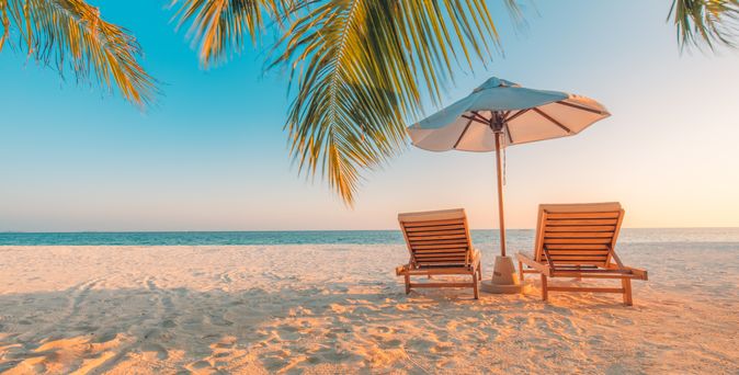 Zwei Liegen mit einem Sonnenschirm stehen unter Palmen am Strand