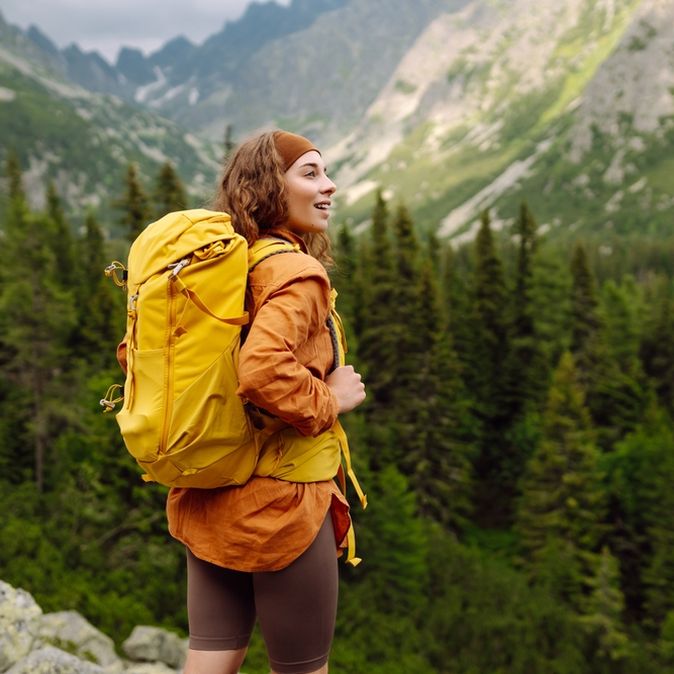 Eine Frau mit gelbem Wanderrucksack steht auf einem Berg und schaut begeistert in die Ferne