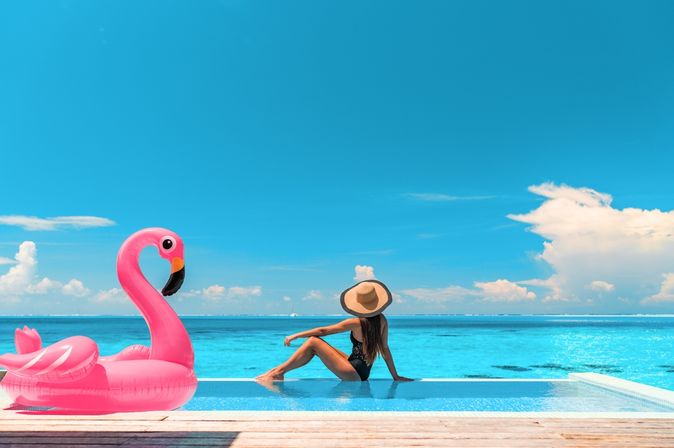 Frau sitzt am Rand eines Pools mit Meerblick, neben Ihr ein aufblasbarer Flamingo