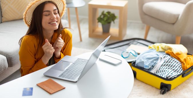 Eine Frau sitzt glücklich vor ihrem Laptop, auf dem Tisch liegen Reisepass und Kreditkarte, im Hintergrund liegt ein gepackter Koffer