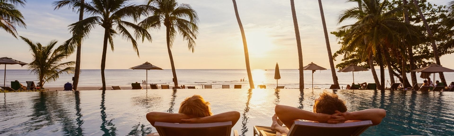 Zwei Personen von hinten fotografiert, Sie liegen auf zwei Liegen auf einem Steg im Pool und schauen auf den Sonnenuntergang über dem Meer