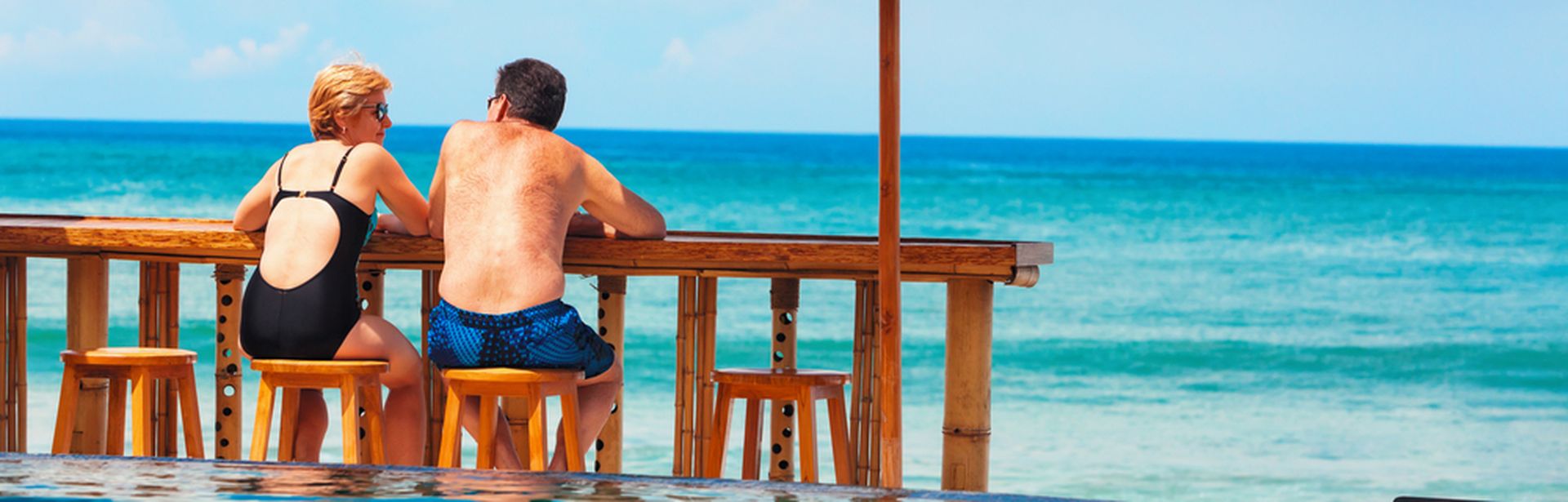 Eine Frau und ein Mann in Badekleidung sitzen an einer Holztheke auf Holzstühlen vor einem Pool und schauen auf das Meer