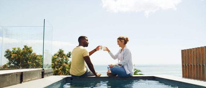 Eine Frau und ein Mann sitzen am Pool und stoßen mit Ihren Getränken an