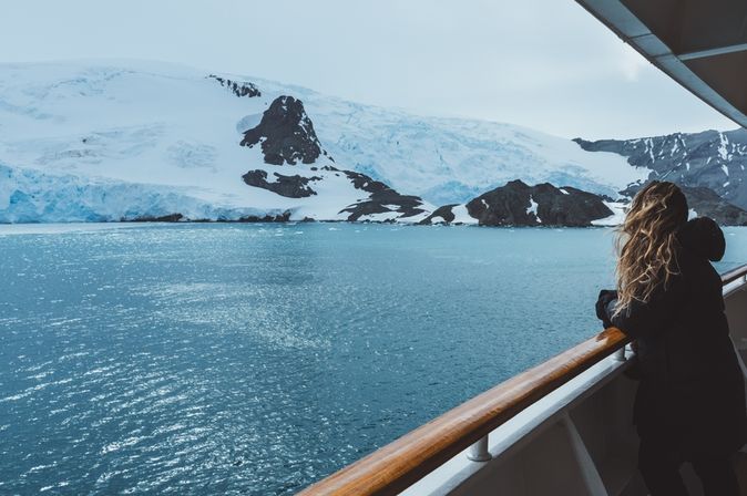 Eine Frau steht mit wehenden Haaren an der Reling eines Kreuzfahrtschiffes und schaut auf schneebedeckte Berge