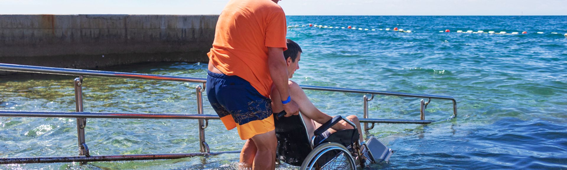 Mann hilft einem anderen Mann im Rollstuhl über eine Treppe ins Meer