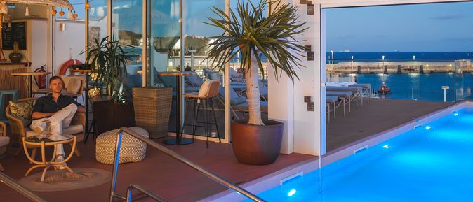 Ein halbüberdachter Pool am Abend, unter der Überdachung stehen Pflanzen und Stühle, auf einem der Stühle sitzt ein Mann