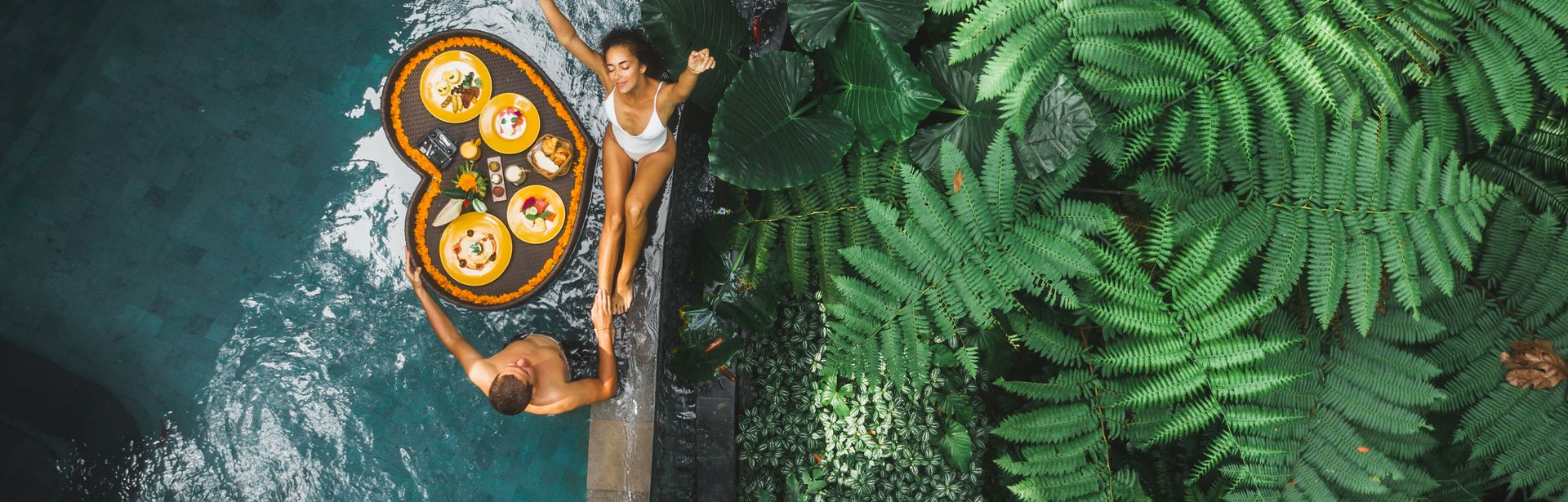 Ein Paar von oben, eine Frau sitzt am Rand eines Pools, ein Mann steht im Pool, zwischen ihnen schwimmt ein Herzförmiges Tablett mit Essen