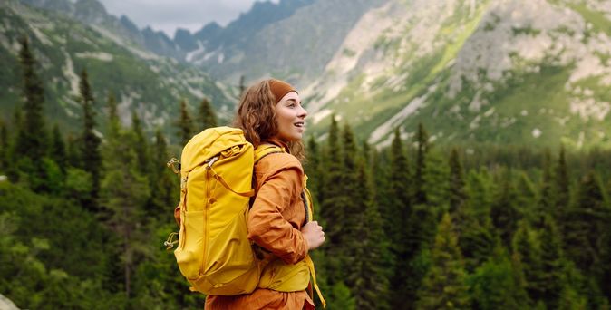 Eine Frau mit gelbem Wanderrucksack steht auf einem Berg und schaut begeistert in die Ferne