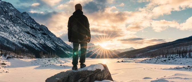 Mann steht leicht erhöht auf einem großen Stein in den Bergen bei Schnee und schaut in die untergehende Sonne