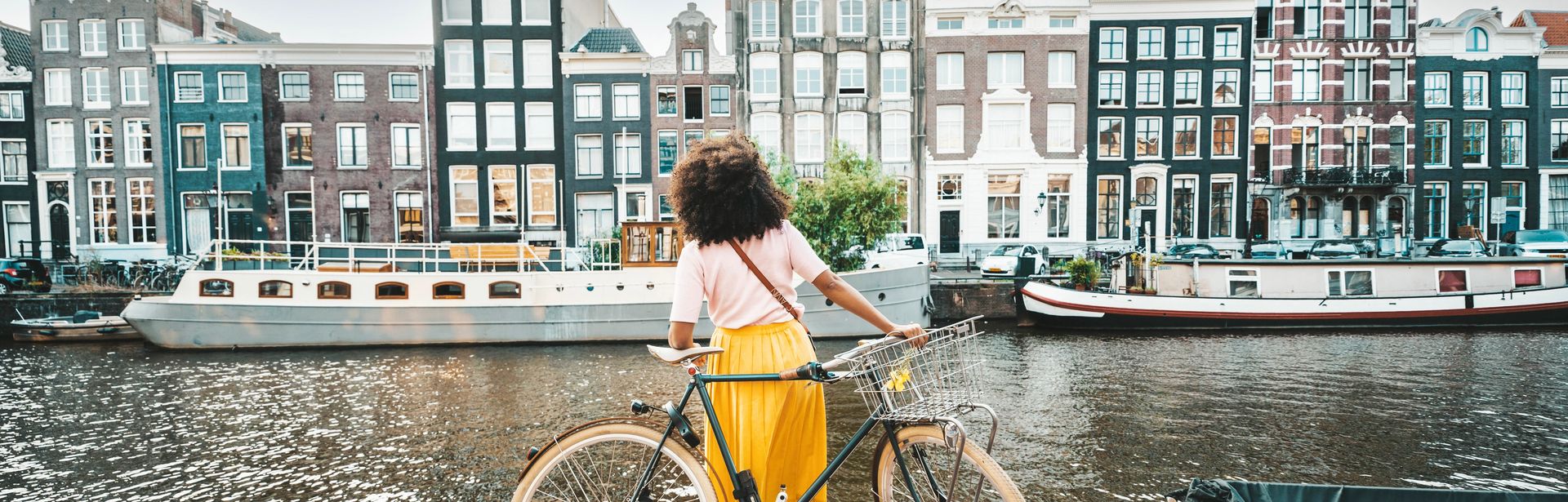 Eine Frau steht mit ihrem Fahrrad vor der Amstel und schaut auf die Häuser auf der gegenüberliegenden Seite