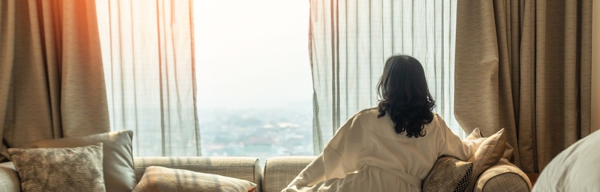 Eine Frau im Bademantel sitzt in einem Hotelzimmer auf dem Sofa und schaut aus den Fenster