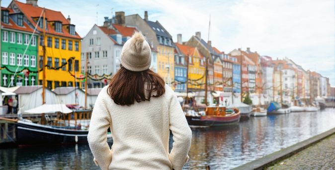 Eine Frau von hinten. Sie trägt eine weiße Jacke und eine Mütze. Sie schaut auf bunte Häuser, das Wasser und einige Boote in Nyhavn in Kopenhagen.