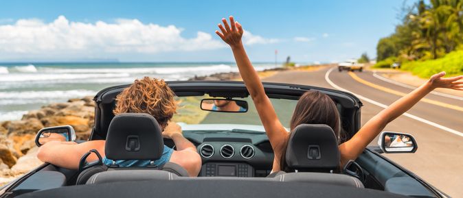 Zwei Personen im Auto mit offenem Verdeck, im Hintergrund kann man das Meer sehen