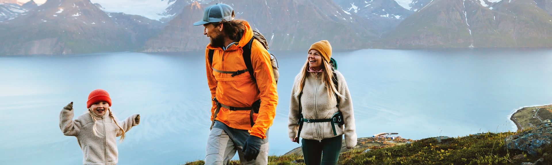 Eine Familie wandert in Winterkleidung an einem Gewässer, im Hintergrund sind verschwommen Berge 
