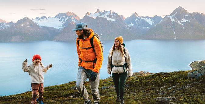 Eine Familie wandert in Winterkleidung an einem Gewässer, im Hintergrund sind verschwommen Berge 