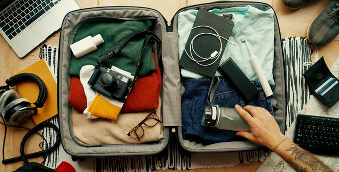 Offener voller Koffer, mit Klamotten, Kamera und einem Ladekabel. Eine Hand packt eine Wasserflasche in den Koffer. Um den Koffer liegen verschiedene Gegenstände herum