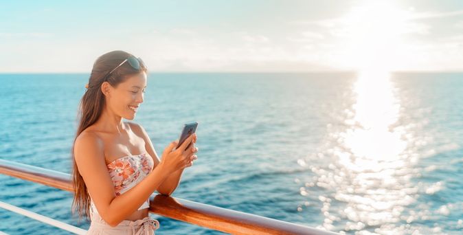 Eine Frau steht lächeln an der Reling eines Kreuzfahrtschiffes und schaut auf ihr Smartphone
