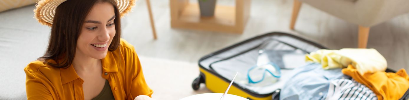 Eine sich freuende Frau tippt an einem Laptop. Im Hintergrund liegt ein gepackter Koffer