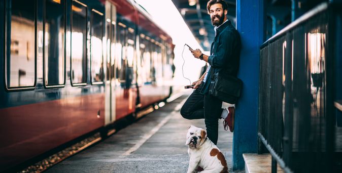 Ein Mann und ein Hund stehen gemeinsam an einem Bahnsteig und schauen in die Kamera