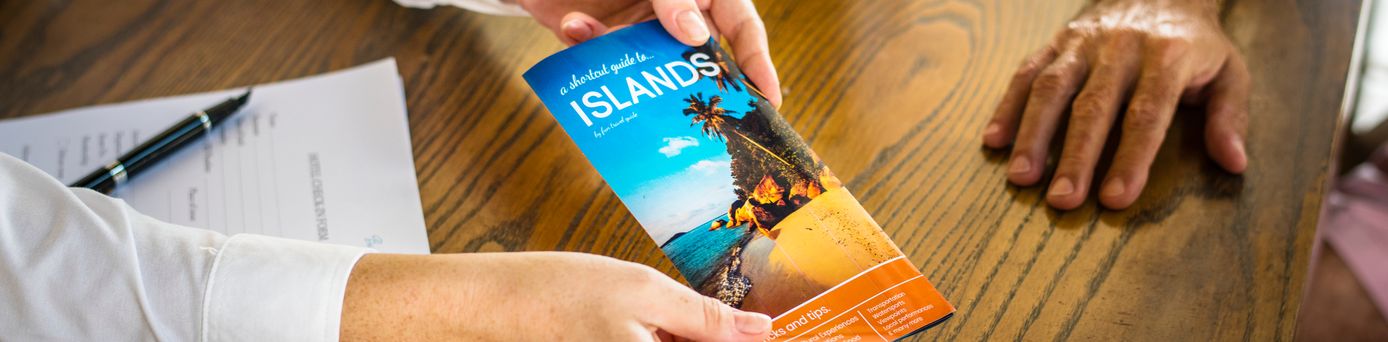 Ein Schreibtisch in einem Reisebüro, der Reiseberater überreicht eine Broschüre zu einem Urlaubsland