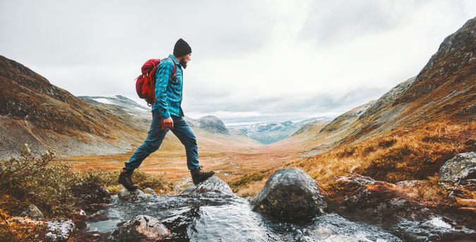 Ein Mann in Wanderkleidung läuft in einer Berglandschaft über Steine in einem Bach