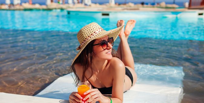 Eine lächelnde Frau im Bikini liegt auf einer Liege in einem Pool und schaut glücklich nach Rechts, in der Hand hält sie einen Orangen Cocktail