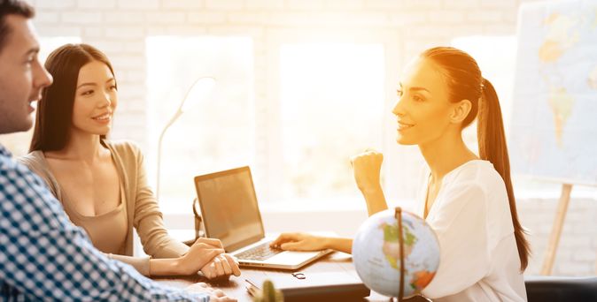 Ein Paar sitzt mit einer Reiseberaterin am Tisch, während das Licht durch das Fenster im Hintergrund den Raum erleuchtet