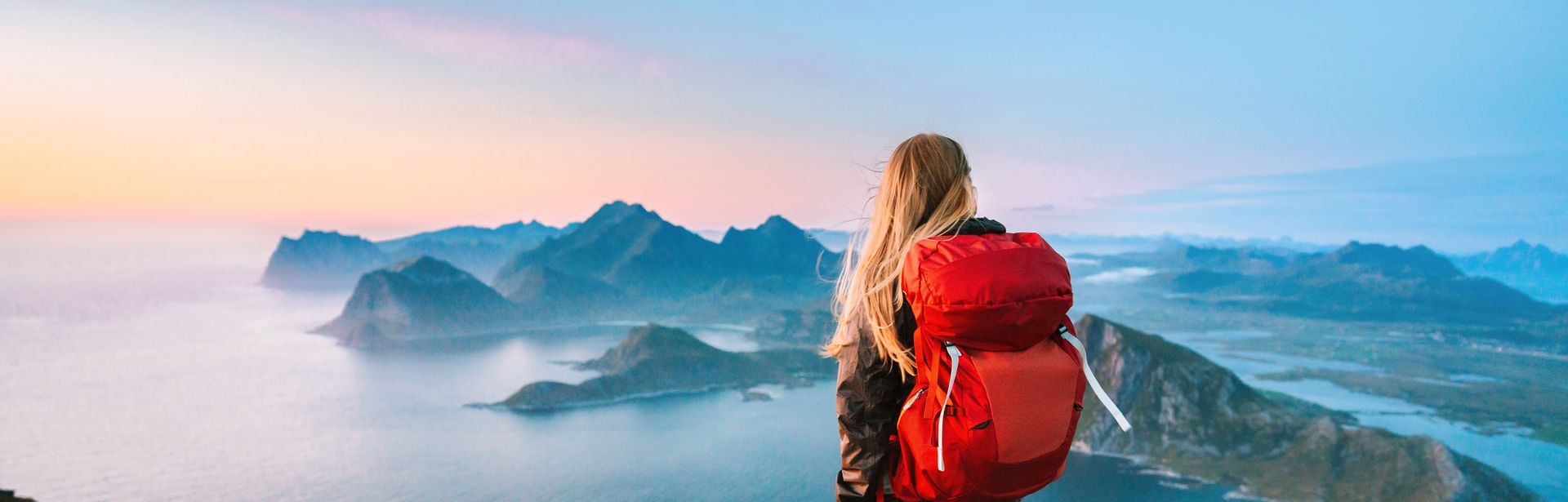 Eine Frau von hinten, Sie trägt einen großen Rucksack und schaut von einem Berg aus den Sonnenaufgang an