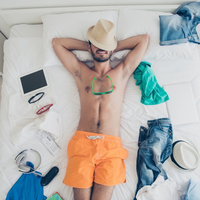 Ein Mann in oranger Badehose liegt auf dem Rücken im Bett, sein Hut liegt auf seinem Gesicht und um ihn herum liegen Anziehsachen, ein Tablet und eine Sonnenbrille