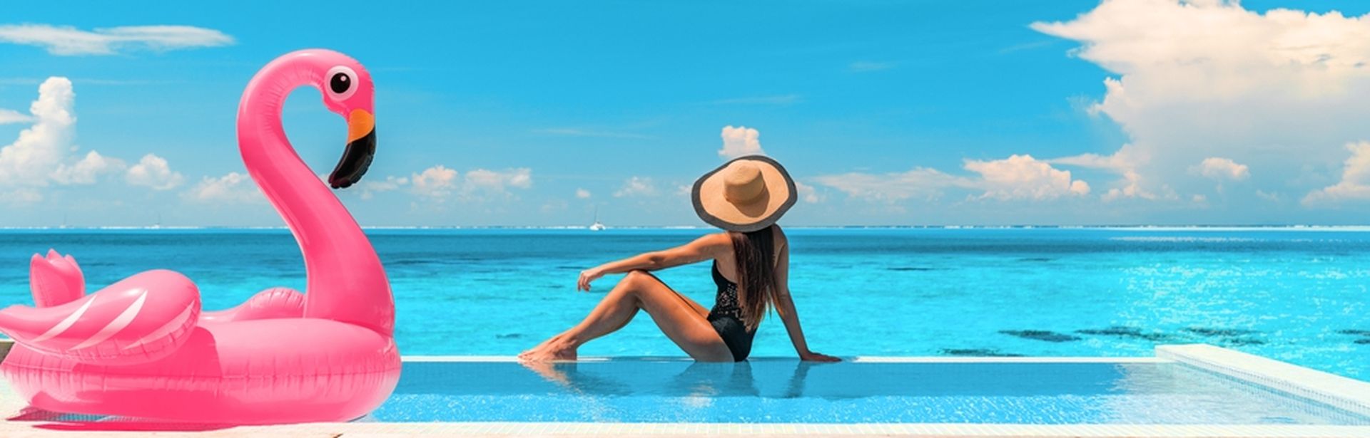 Frau sitzt am Rand eines Pools mit Meerblick, neben Ihr ein aufblasbarer Flamingo