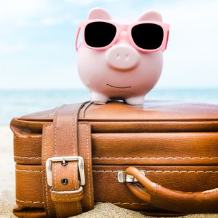 Ein rosanes Sparschwein mit Sonnenbrille sitzt auf einem Koffer am Strand